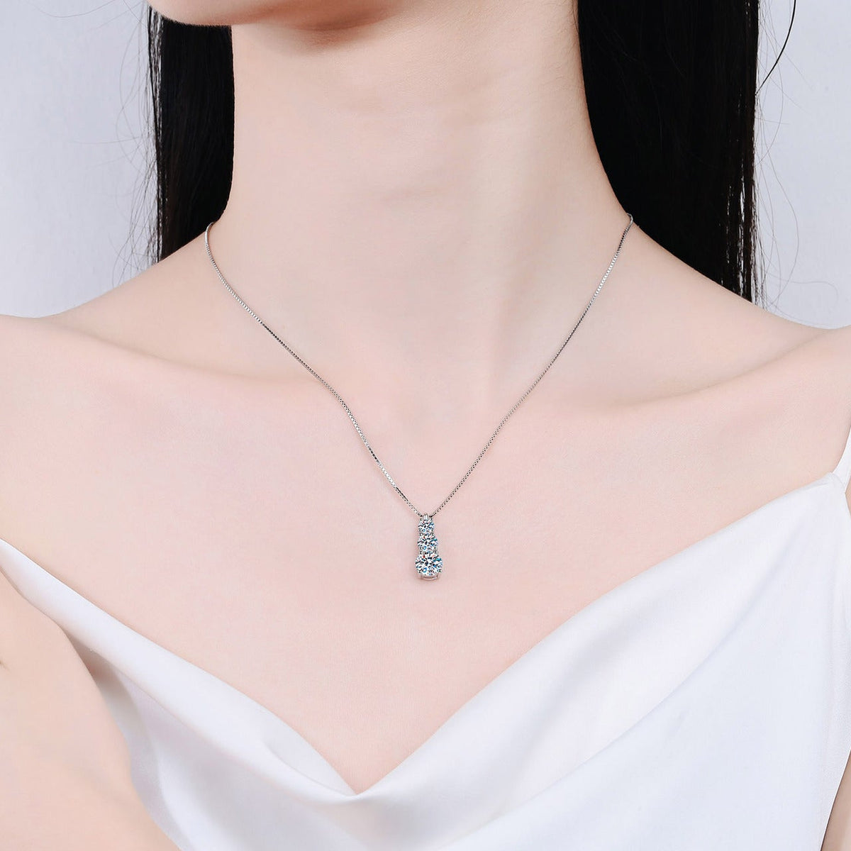 A close-up of a model wearing a delicate silver necklace with a small, clear-stone pendant, styled with a white camisole top against a light gray background.