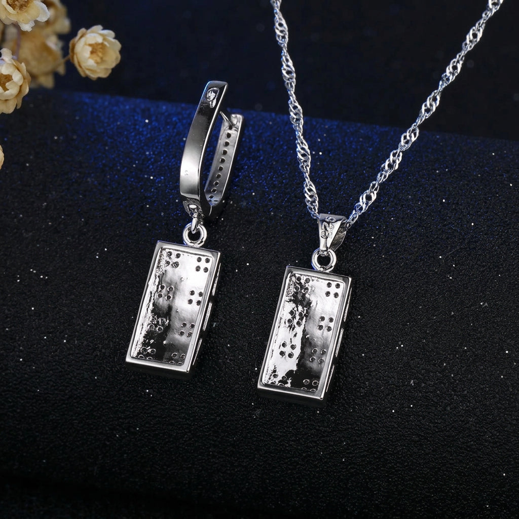 A silver-toned jewelry set featuring a rectangular pendant necklace and matching huggie hoop earrings, both with a textured, metallic dot pattern, displayed on a dark textured background. The brand logo 'Jexxi' is visible in the top left corner.