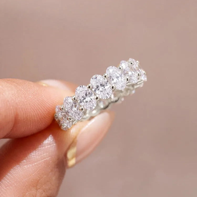 A close-up of a diamond eternity ring held between fingers against a soft, warm beige background. The ring features a continuous row of round brilliant-cut diamonds set in a silver-colored metal band.