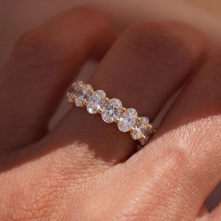 A close-up of a gold eternity ring with round clear gemstones worn on a finger.