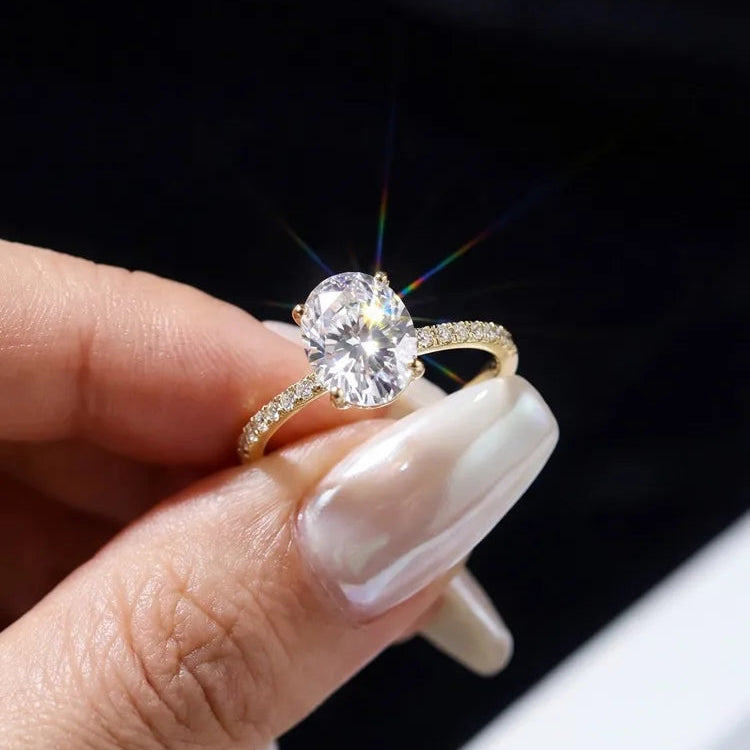 A close-up of a hand with long, glossy nude manicured nails holding a gold ring with a large round brilliant-cut clear gemstone and a pavé band.