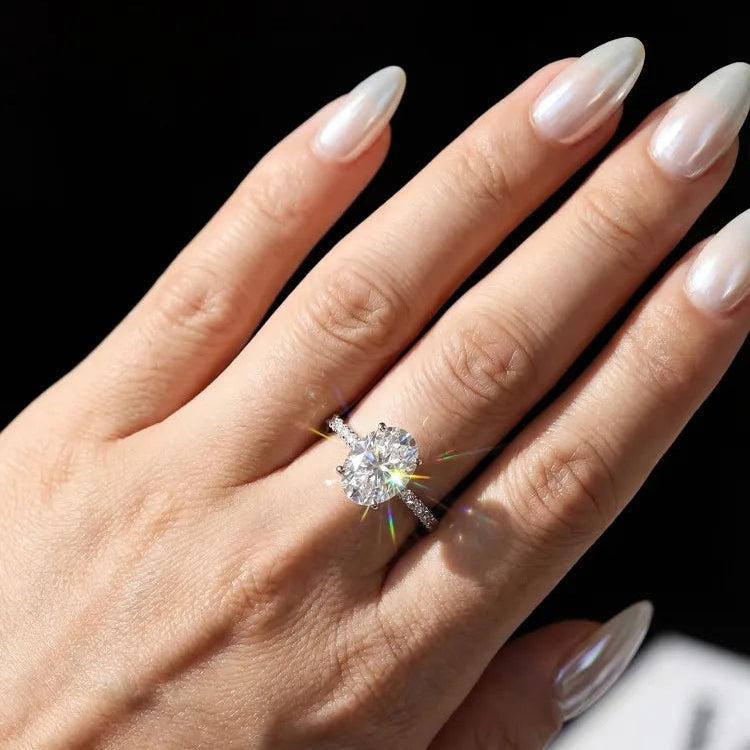 A close-up of a hand with long, glossy nude manicured nails wearing a sparkling diamond engagement ring. The ring features a large round brilliant-cut center stone set in a silver-toned band with small pavé accents.