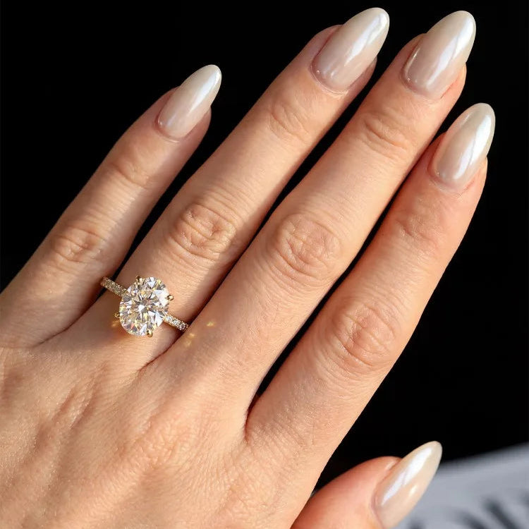 A close-up of a hand with long, glossy nude manicured nails wearing a yellow gold ring with a large round brilliant-cut diamond and smaller accent diamonds.