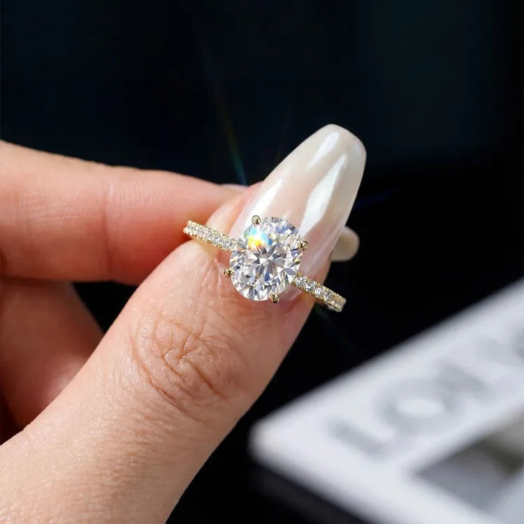 A close-up of a hand with long white manicured nails holding a gold ring with a large round clear gemstone and small side stones, against a dark blurred background.
