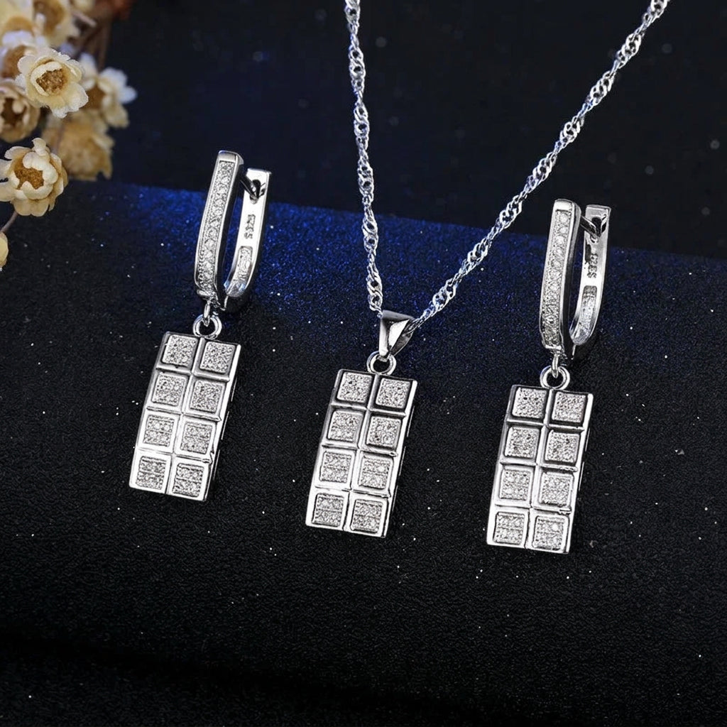 A jewelry set featuring a silver-tone necklace and matching earrings with rectangular pendant charms detailed with small clear stones, displayed on a dark textured surface with a decorative floral corner and a visible JEXXI logo.