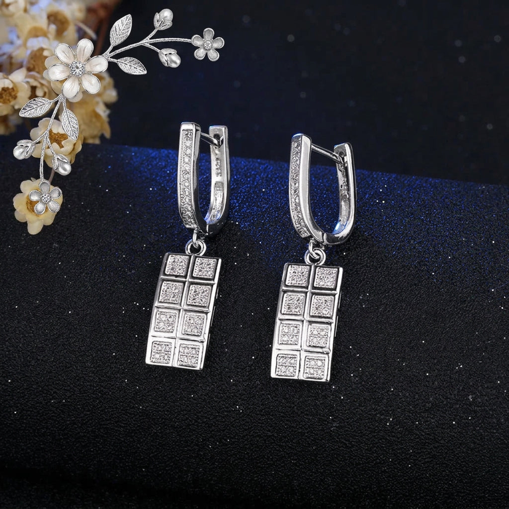 A pair of silver-toned drop earrings with rectangular grid charms set with clear stones, displayed on a dark textured surface with a decorative floral corner and a visible JEXXI logo.