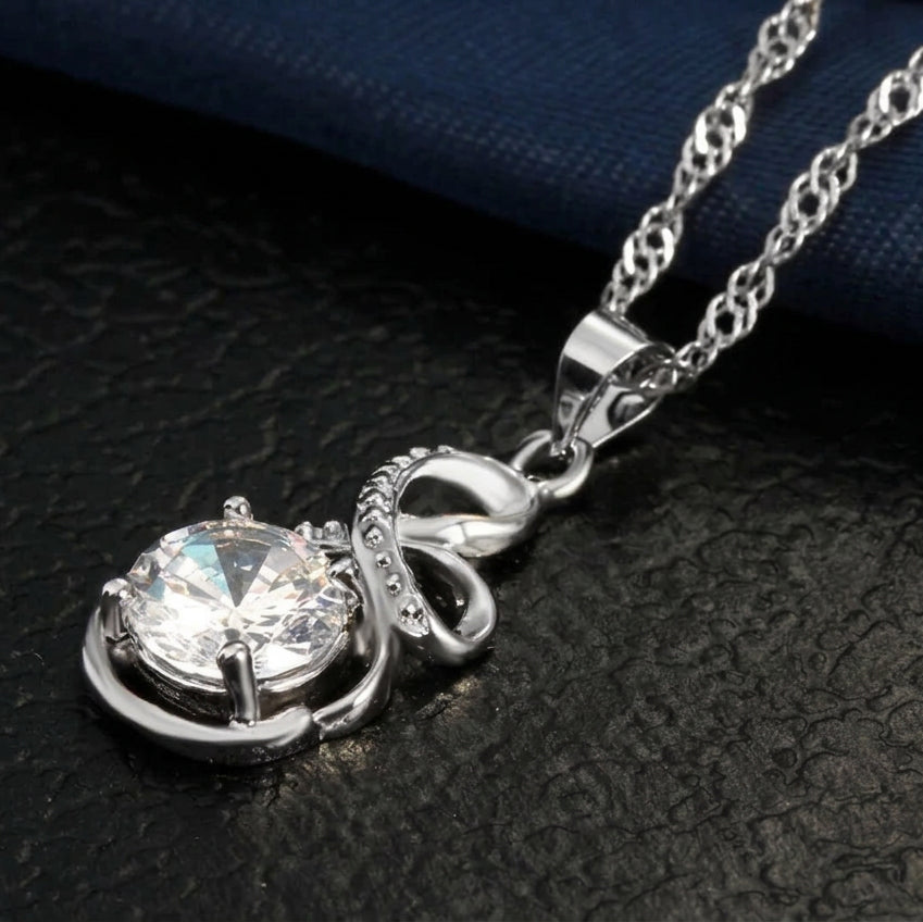A silver-tone pendant necklace featuring a large round clear gemstone in a decorative setting, suspended from a matching chain on a dark textured surface.