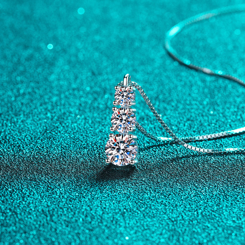 A silver-toned necklace with a three-stone diamond pendant, displayed on a sparkling teal textured surface.