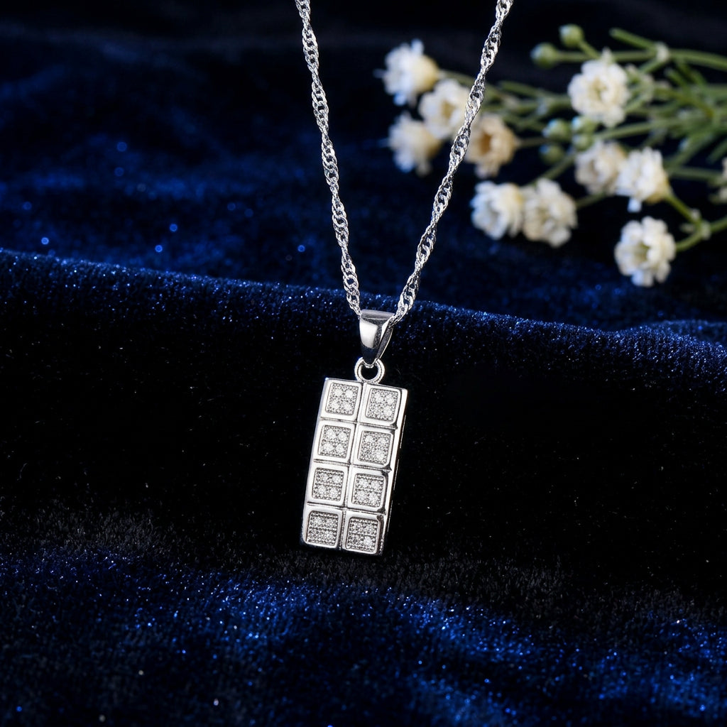 A silver necklace with a rectangular pendant designed to look like a chocolate bar, featuring small clear stones in each section. The necklace is displayed on a dark textured background with a blue fabric accent, and a brand logo 'Jexxi' is visible in the top left corner.