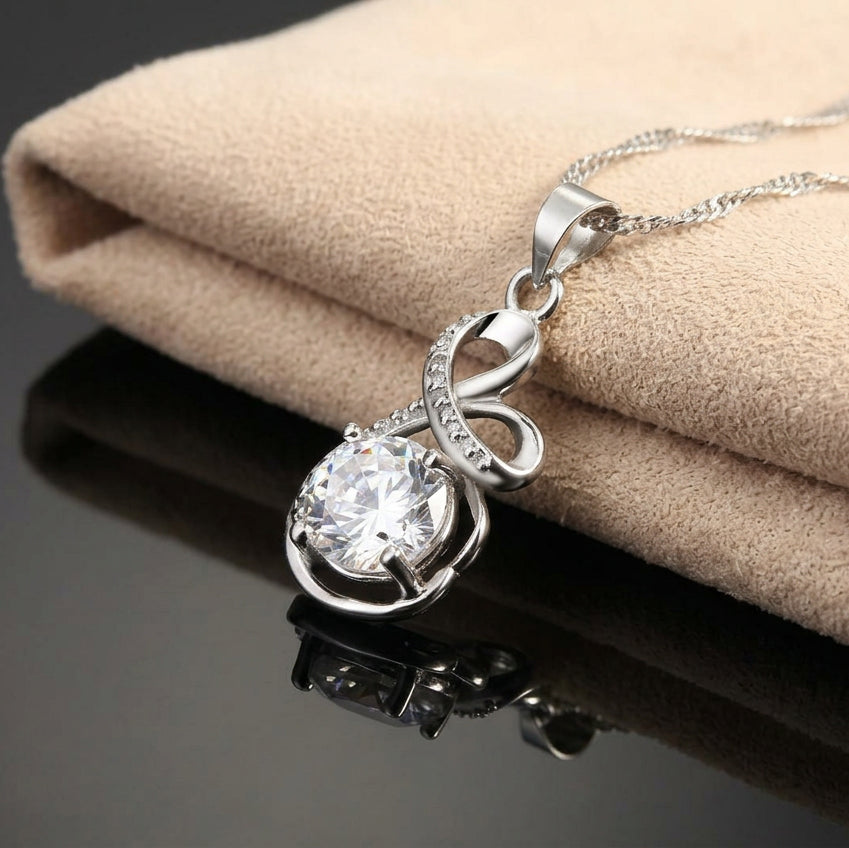 A silver necklace with a round clear gemstone pendant, resting on a textured beige fabric surface with a black reflective surface below.
