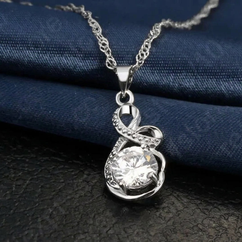 A silver pendant necklace with a clear round gemstone and small accent stones, resting on a dark textured surface with a folded navy blue fabric backdrop.