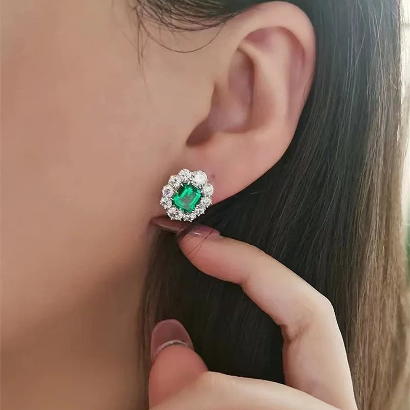A close-up of a person's ear wearing a green rectangular stud earring with a halo of small clear stones, with fingers gently touching the side of the head.