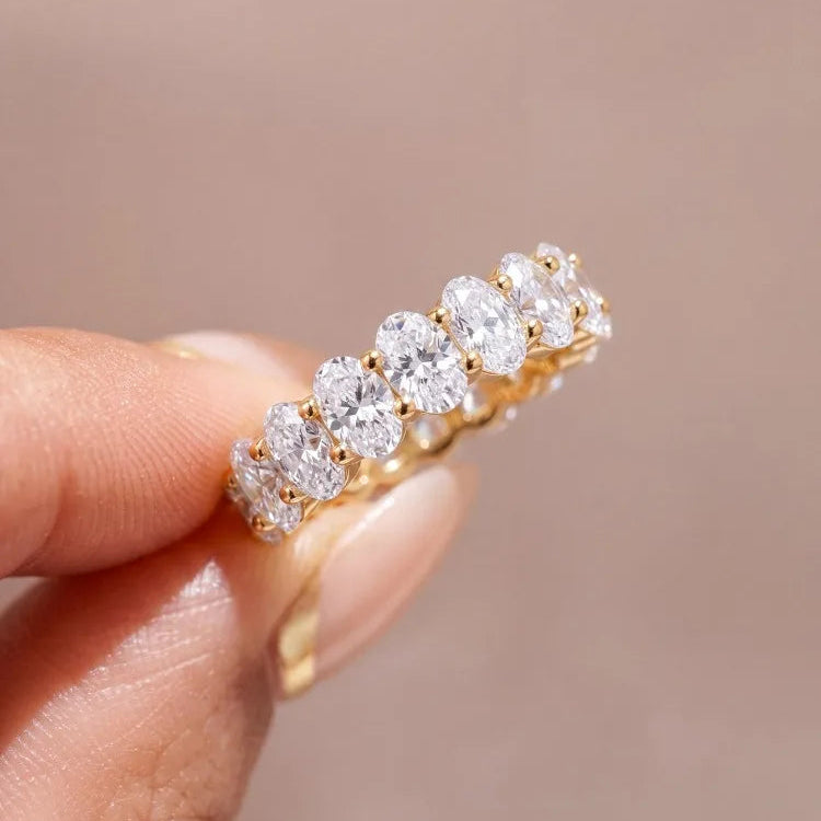 A close-up of a hand holding a yellow gold eternity ring set with round brilliant-cut diamonds against a soft, warm beige background.